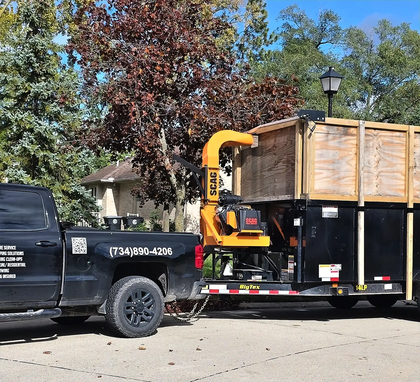 Black pickup truck hitched to a trailer carrying an orange wood chipper and a wooden-sided box, parked on a driveway in front of a brick house and autumn trees